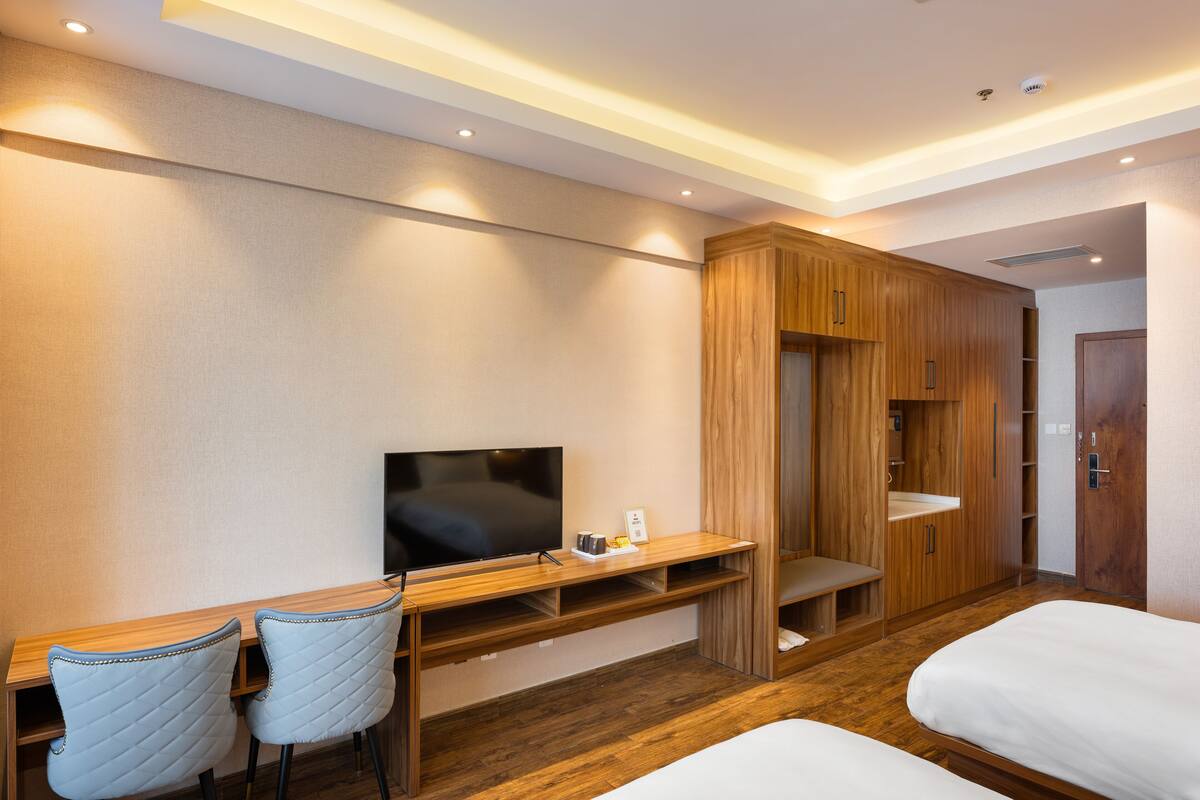 Ein Hotelzimmer mit Schreibtisch aus Holz, zwei Stühlen, einem Flachbildfernseher, einem Kleiderschrank und zwei Betten.