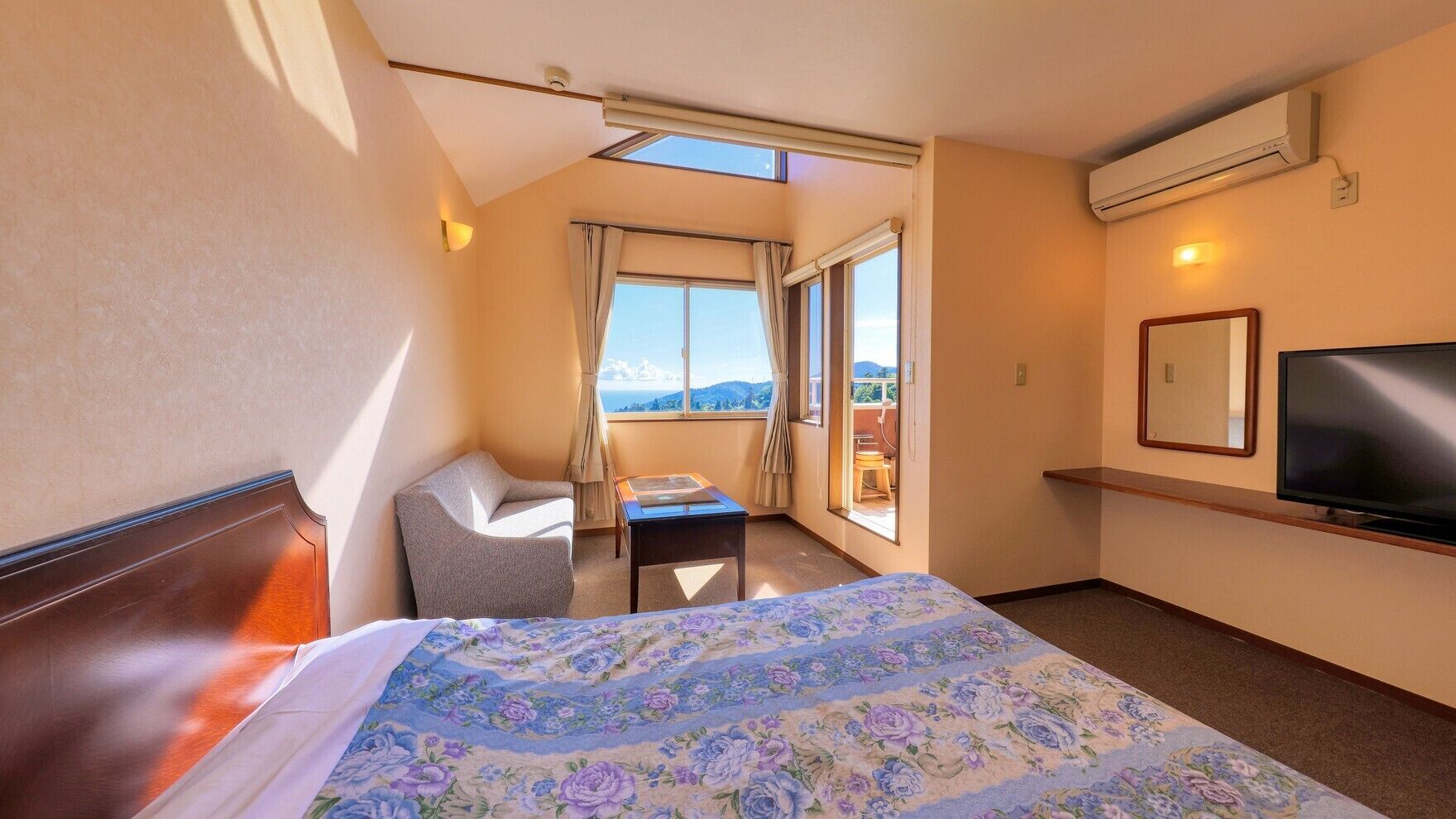 A hotel room with a bed, a chair, a small table, a TV, and a balcony with a view.