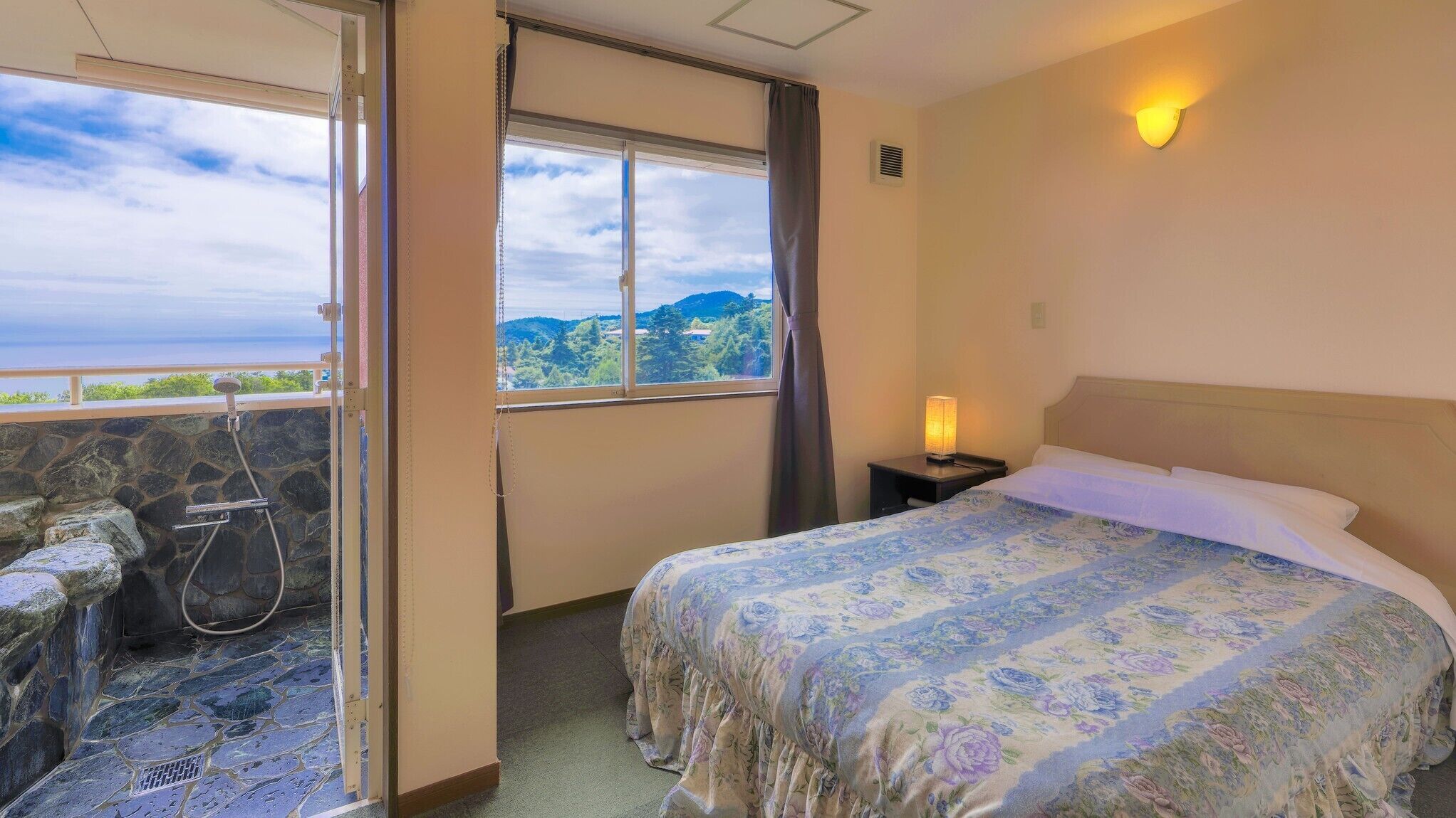 A hotel room with a bed, a nightstand, a lamp, a shower area, and a balcony with a view.