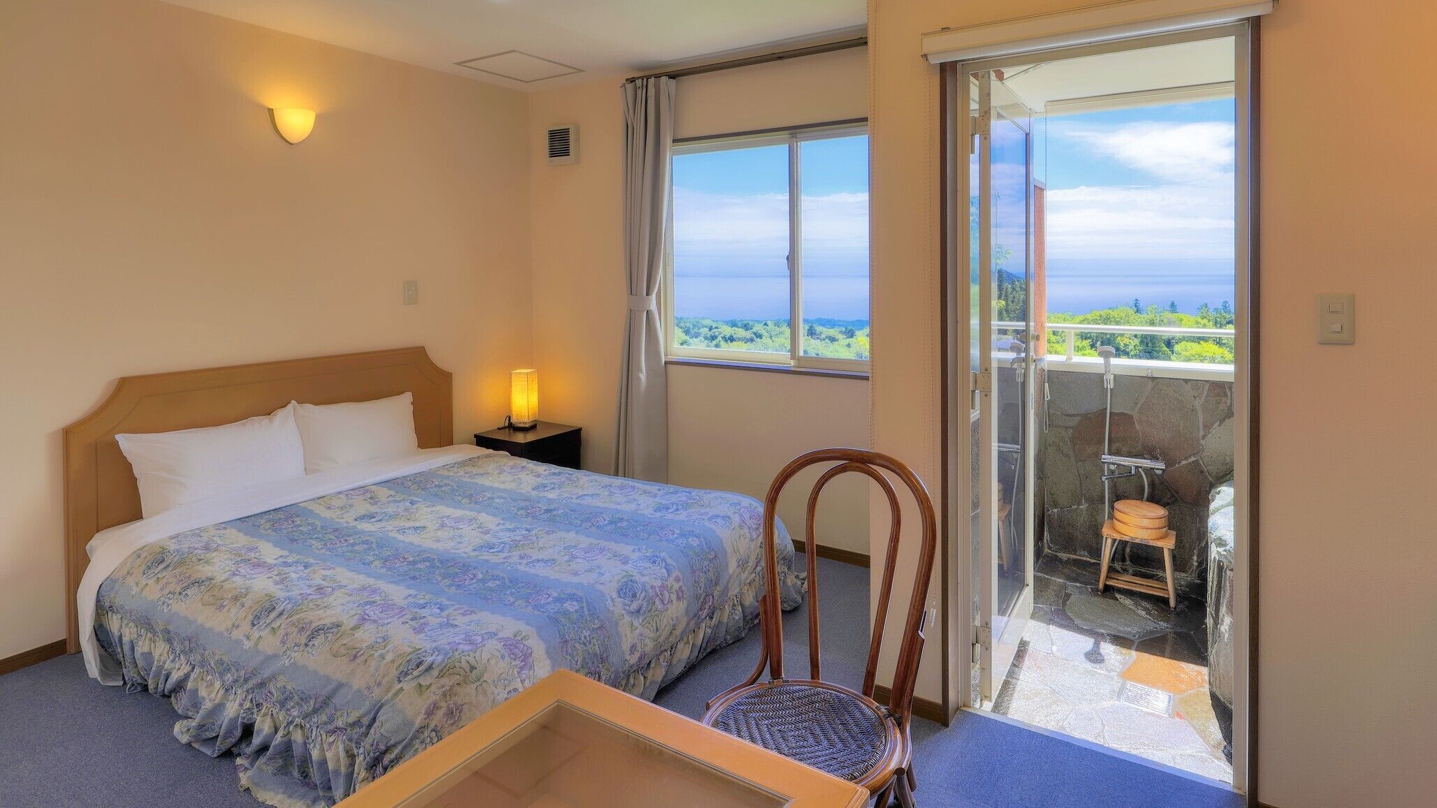 A bedroom with a bed, a nightstand, a lamp, a chair, and a balcony with a view.