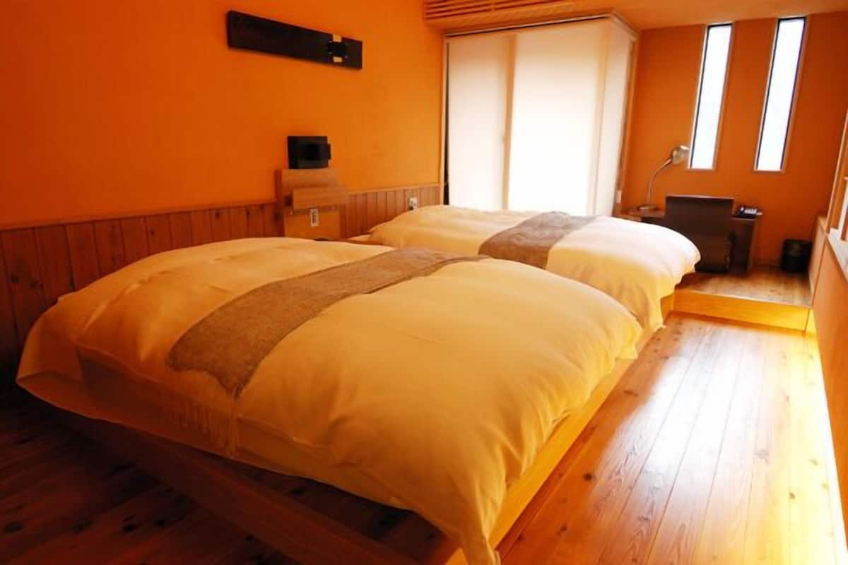 A room with two beds, a wooden floor, a window, and a desk with a lamp.