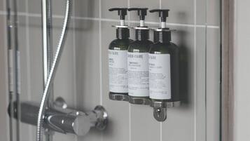 Zona de ducha con puerta de cristal y ducha de mano. Tres botellas de productos para el cabello están fijadas a la pared.