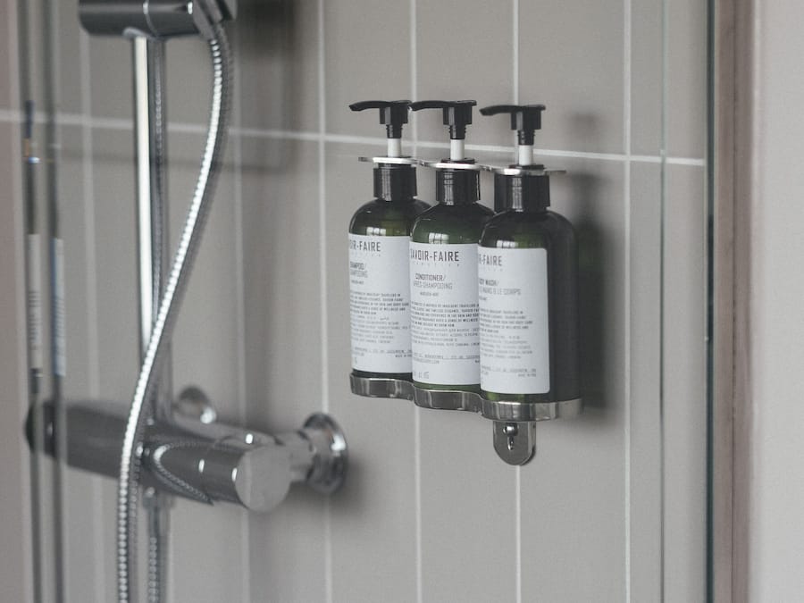 Zona de ducha con puerta de cristal y ducha de mano. Tres botellas de productos para el cabello están fijadas a la pared.