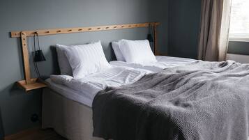 Un lit bien fait, avec du linge de lit blanc et une couverture tricotée grise, une tête de lit en bois et une table de chevet avec une lampe.