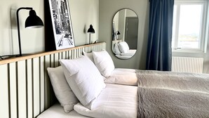 Una cama bien hecha con sábanas blancas y un plaid texturizado, un espejo redondo, dos lámparas de escritorio negras y una fotografía en blanco y negro en la pared.