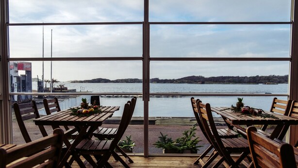 Una terraza de madera con mesas y sillas orientada a un amplio ventanal con vistas a un cuerpo de agua y colinas distantes.