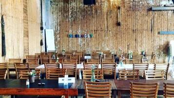 A rustic dining area with wooden tables and chairs, a stage with a banner, and a rustic wall.