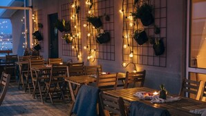 Un espacio de comedor al aire libre con mesas y sillas de madera, luces colgantes y macetas suspendidas.