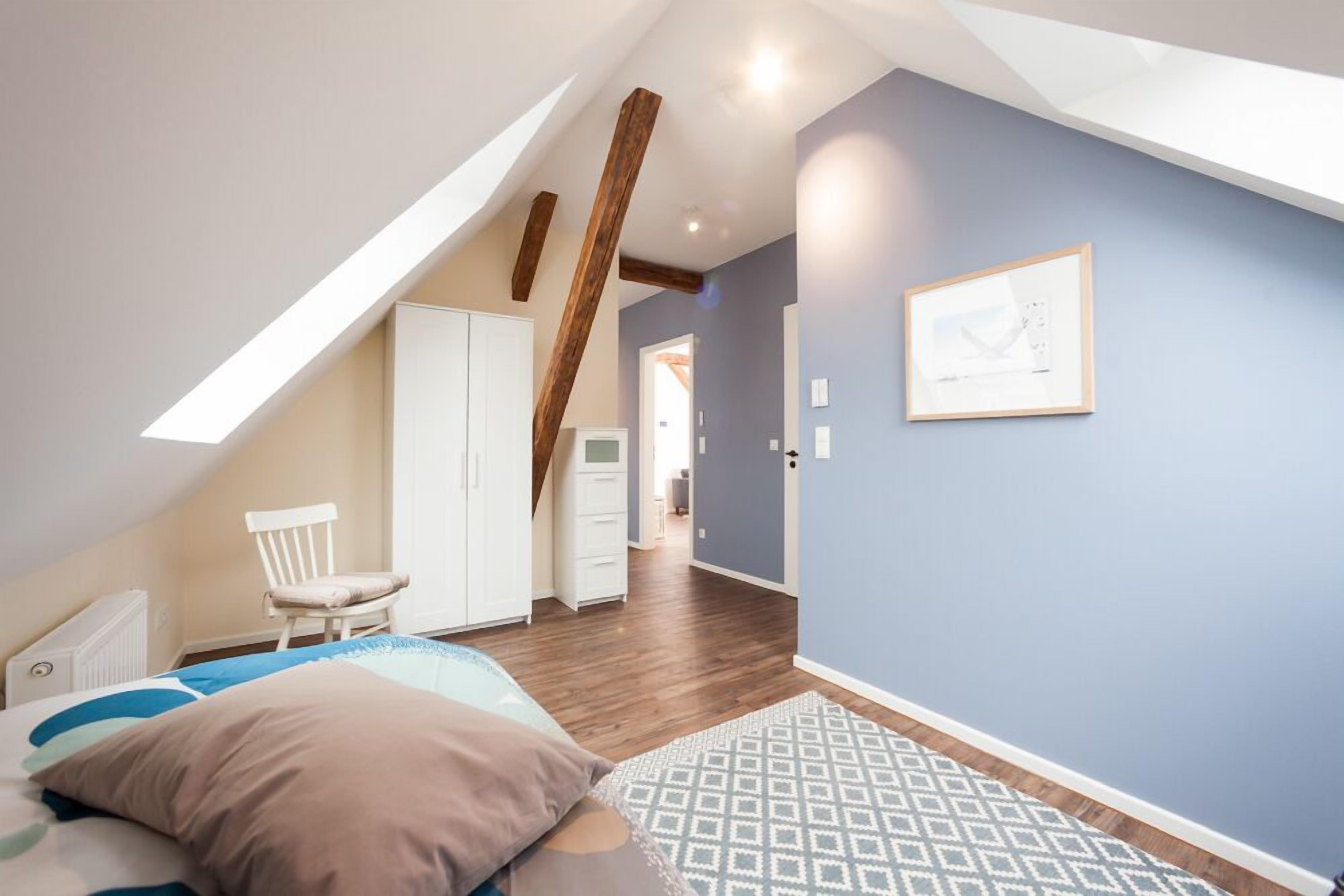 Ein Schlafzimmer mit geneigter Decke, Holzbalken, einem Bett mit Kissen, einem Sessel, einem Kleiderschrank und einem gerahmten Bild an der Wand.