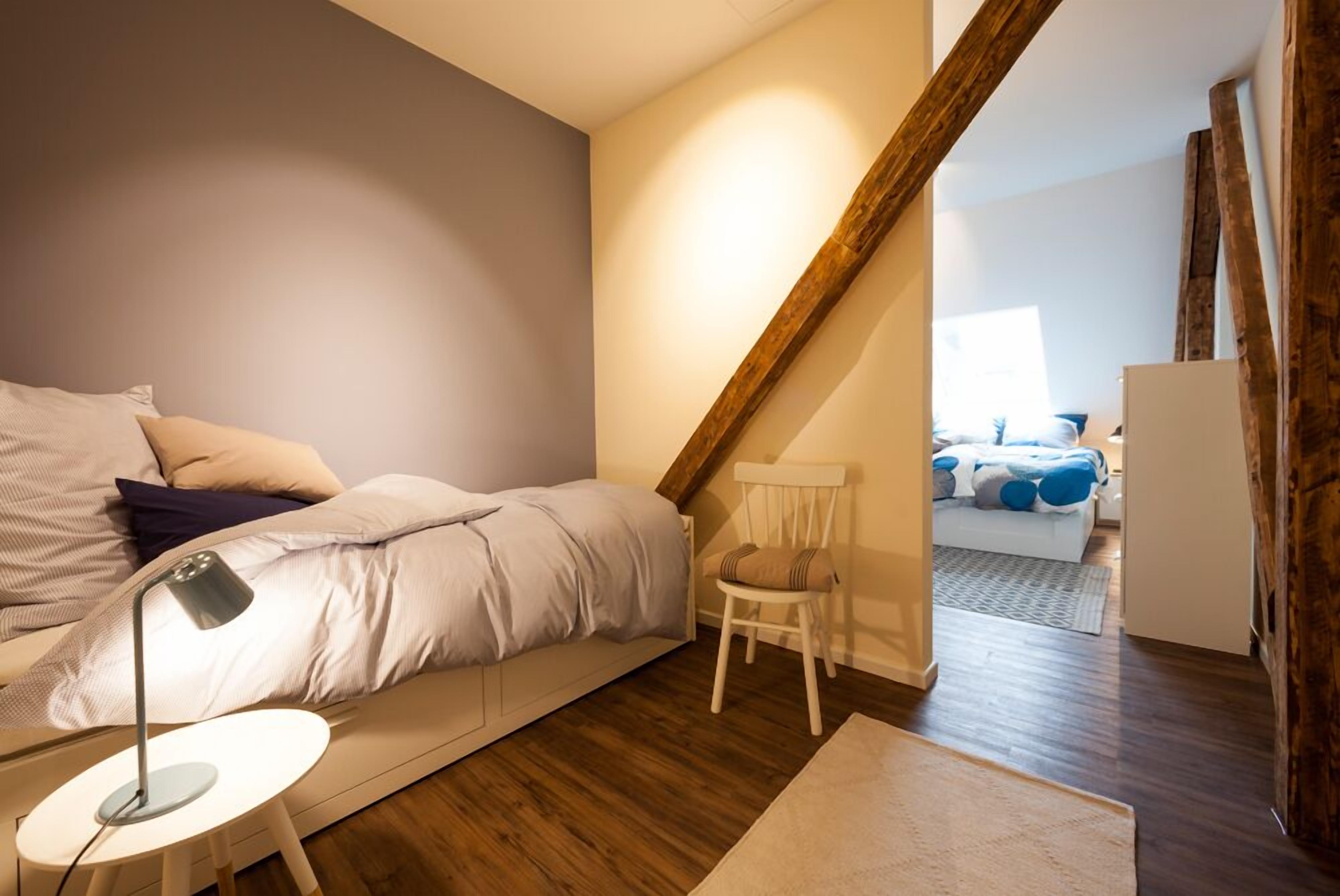 A bedroom with a loft bed, a bedside table with a lamp, a chair, and a view into another room with a bed and a dresser.