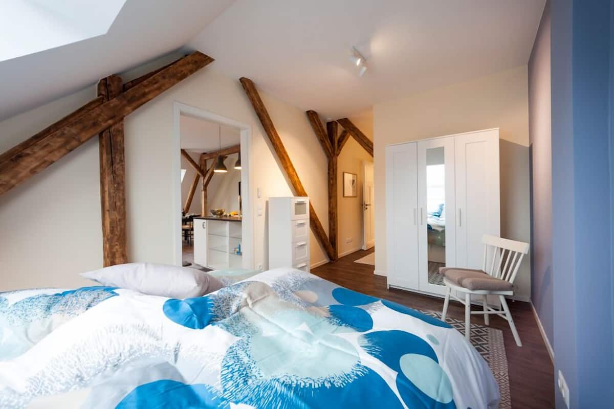 A bedroom with a sloped ceiling, wooden beams, a bed with a blue and white patterned bedspread, a white wardrobe, and a chair.