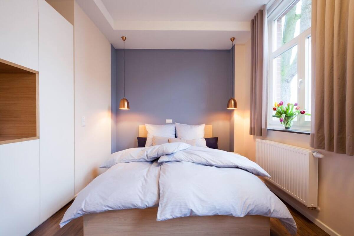 A neatly made bed with white linens, two bedside lamps, a window with curtains, and a radiator.