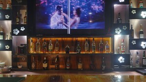 Zona de bar con una pantalla de televisión mostrando una pareja, estantes con botellas de licor y diseños florales decorativos en las paredes.