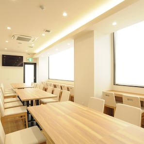 A conference room with long tables and chairs, a projector screen, and a flat-screen TV on the wall.