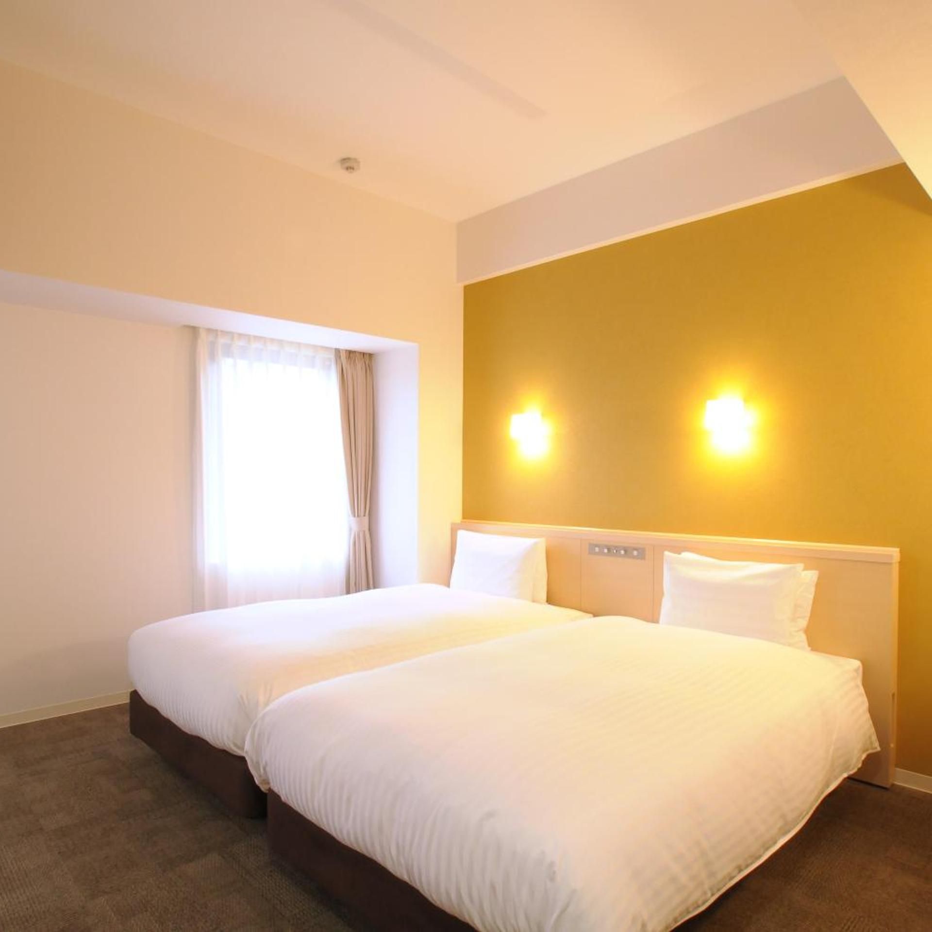 A hotel room with two beds, a window with curtains, and wall-mounted lights.