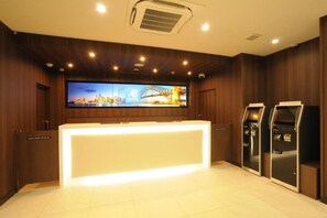 A hotel reception area with a reception desk, a large flat-screen TV displaying a cityscape, and two self-service kiosks.