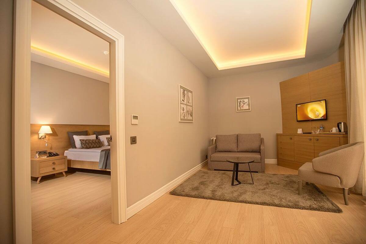 A modern hotel room with a neatly made bed, a sitting area with a sofa and armchair, a small table, and a TV mounted on the wall.