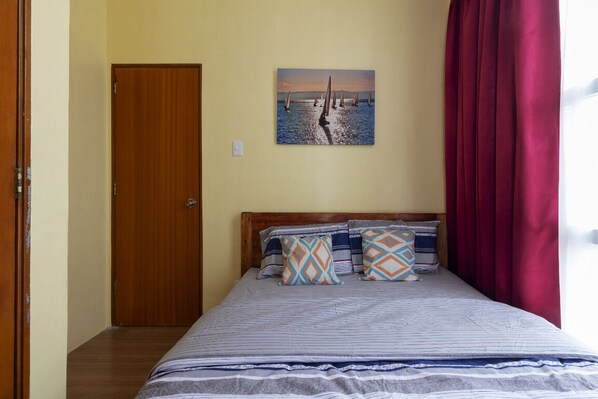 Un dormitorio con una cama, una puerta, un cuadro y una ventana con cortinas.