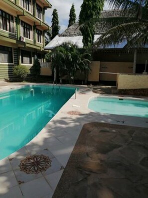 A swimming pool with a tiled deck, surrounded by a building with multiple windows and a palm tree. - Diani Gift Guest House (Diani Beach)