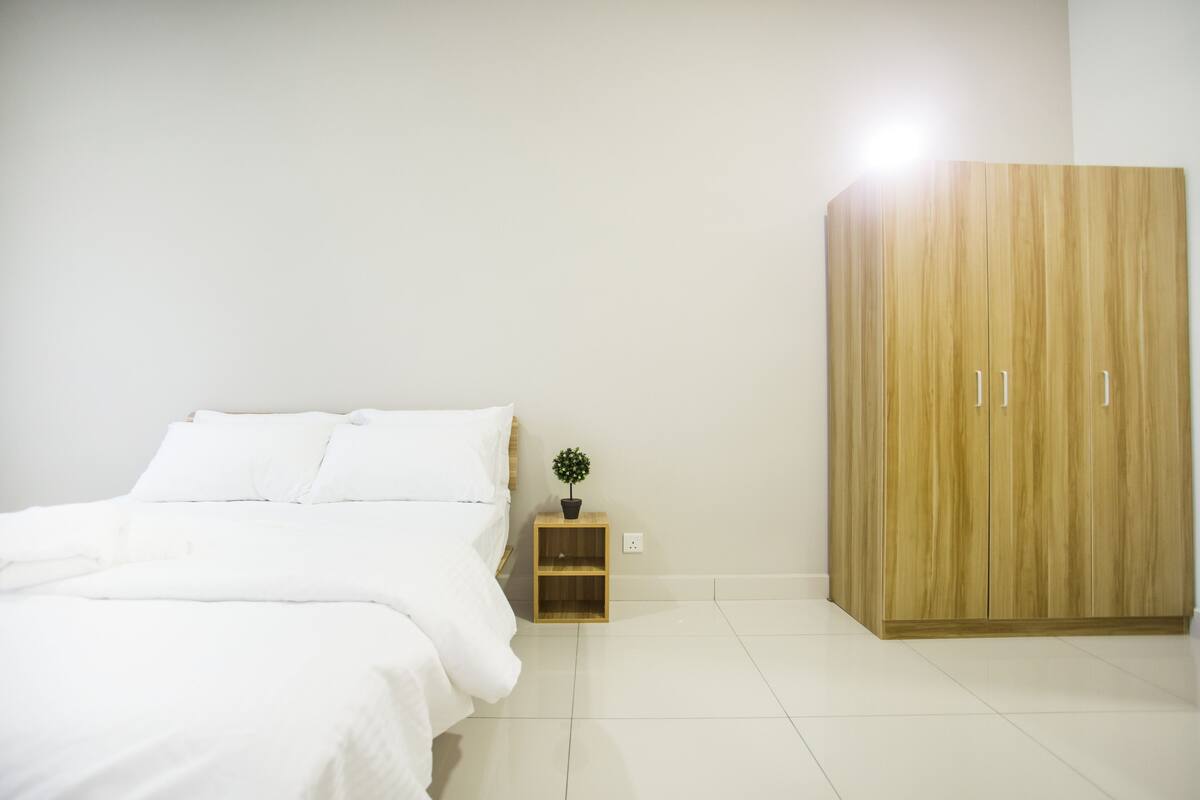 Une chambre à coucher avec un lit, une armoire en bois, une petite table de chevet en bois ornée d’une plante, et un mur blanc.