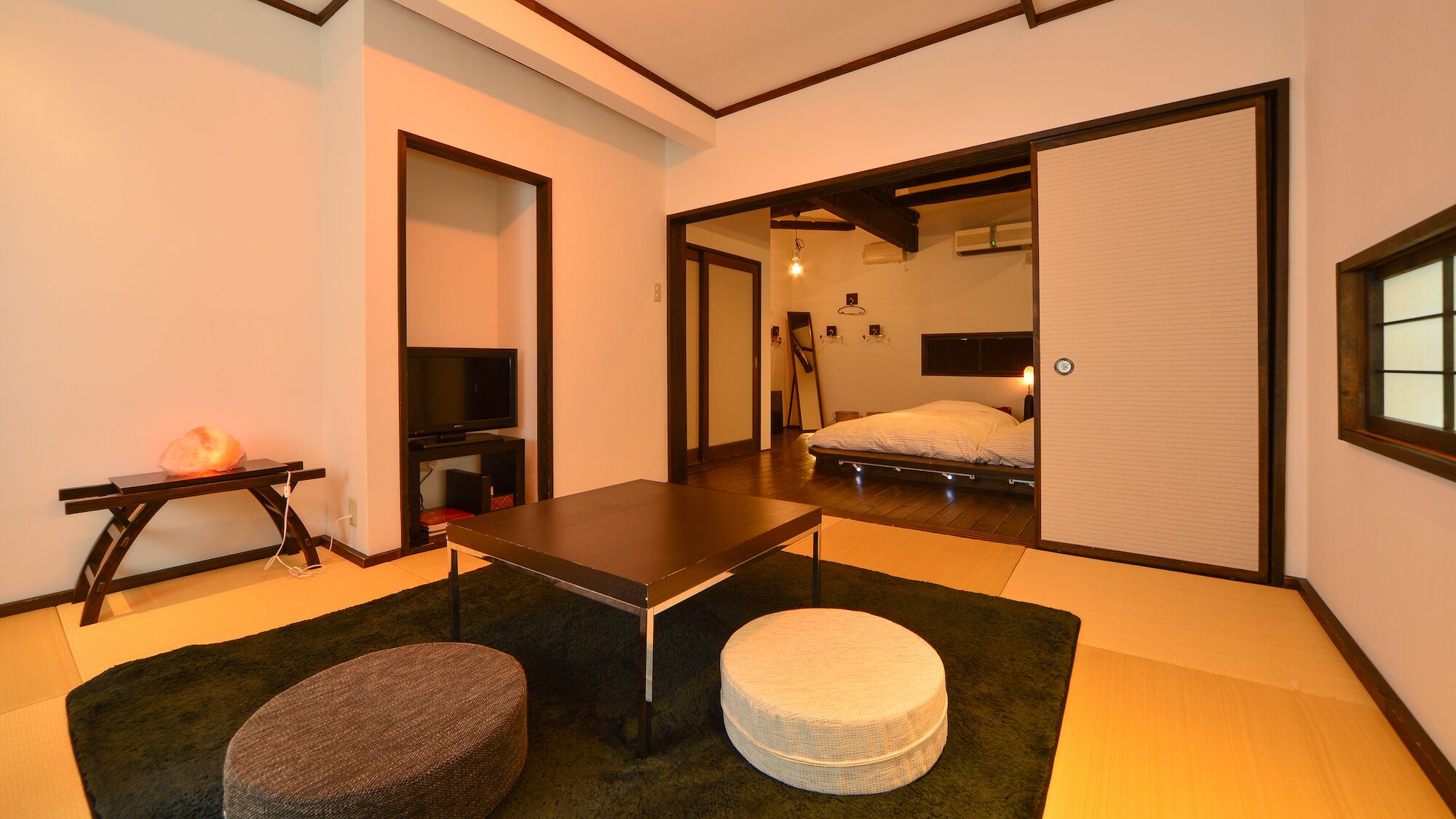 Ein traditioneller japanischer Raum mit Tatami-Boden, einem niedrigen Tisch, einem Futonbett, einem Fernseher und einer verschiebbaren Tür zu einem anderen Raum.