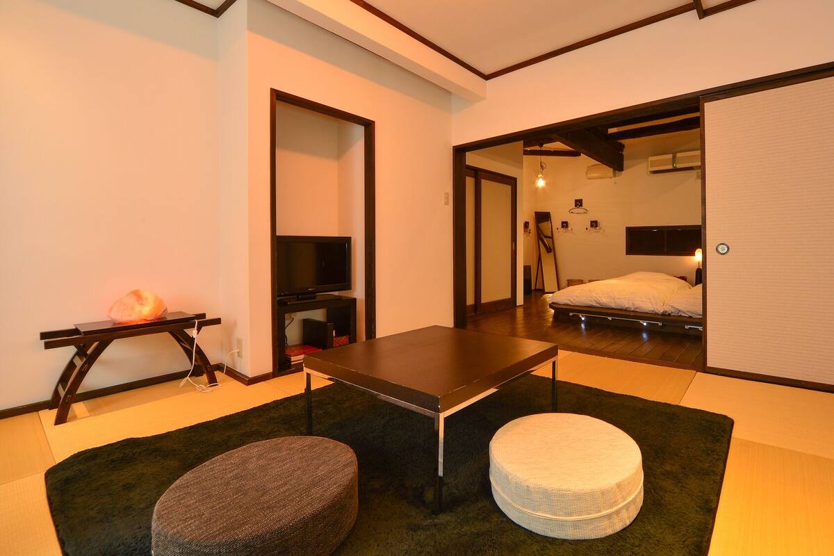 A traditional Japanese-style room with tatami flooring, a low table, a futon bed, a television, and a sliding door leading to another room.