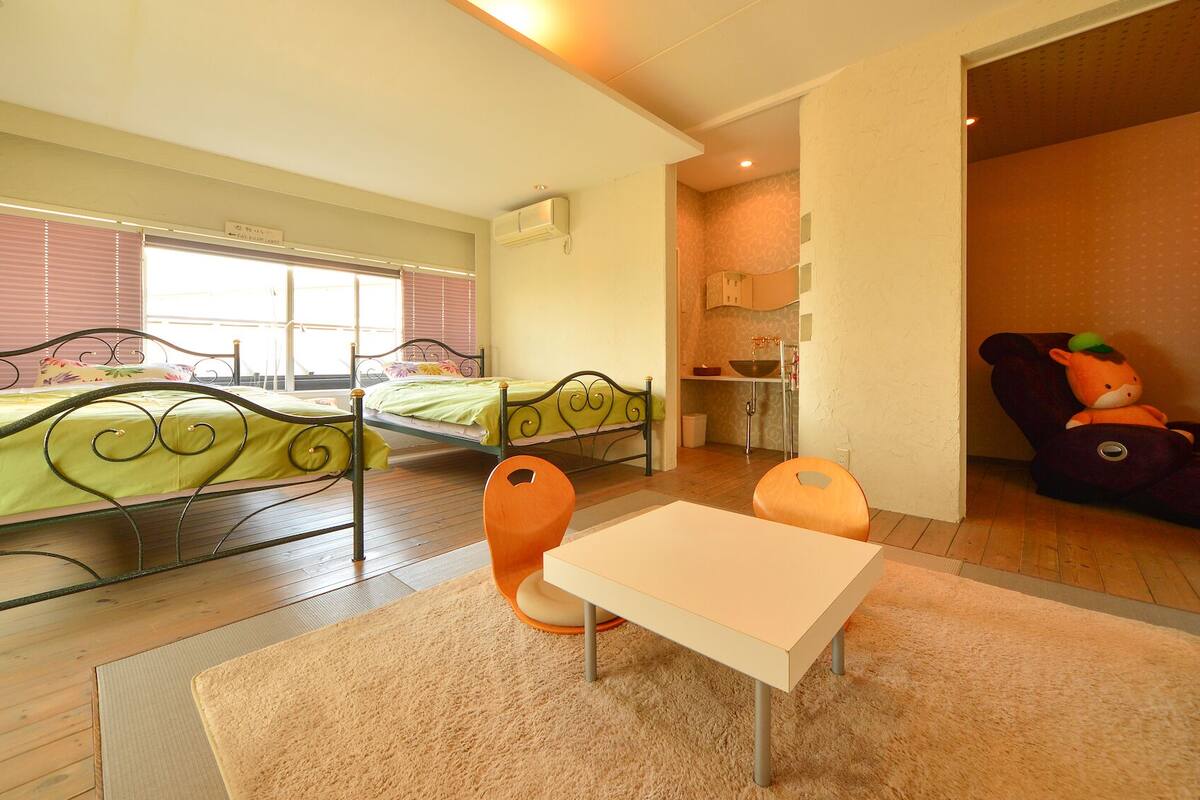 A room with two beds, a small table, and a chair. There is a kitchenette area with a sink and a chair.