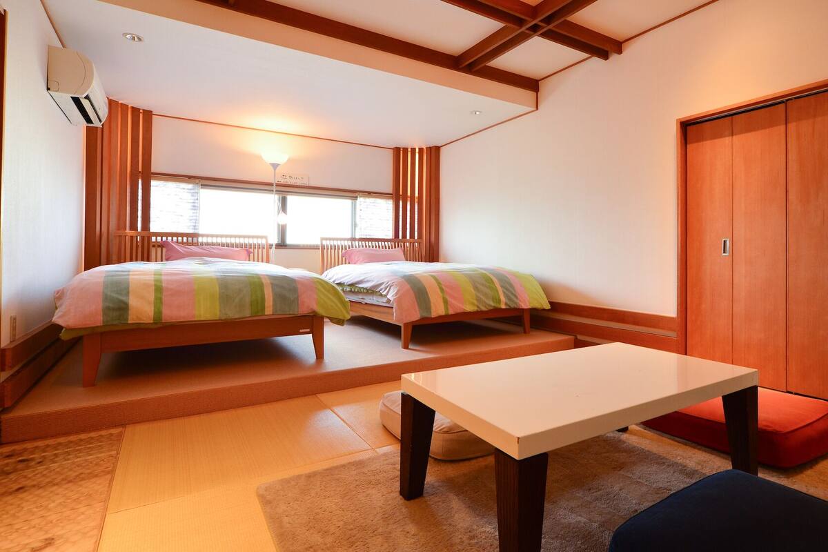 A room with two beds, a table, a wardrobe, and a window with blinds.