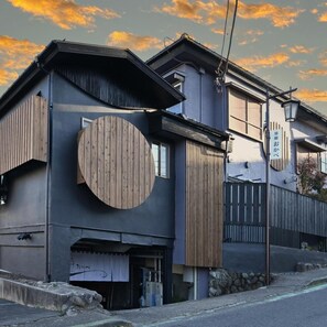 A modern two-story building with a large circular wooden panel on the facade, a small entrance with a sign, and a street lamp. - Ikaho Onsen Akari No Yado Okabe (Shibukawa)
