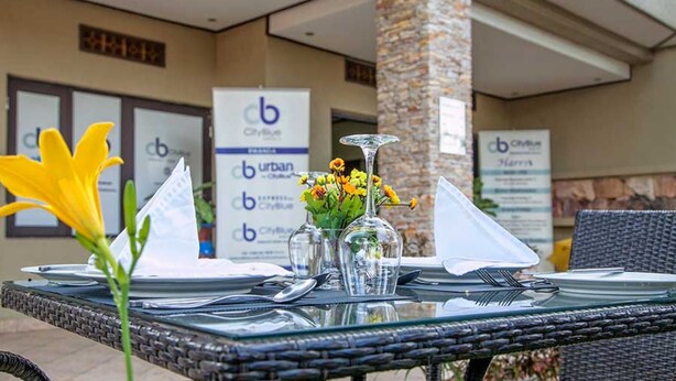 Un área de comedor al aire libre con una mesa dispuesta para dos personas, que incluye un jarrón de vidrio transparente con flores amarillas, una botella de vidrio transparente y una servilleta blanca doblada.