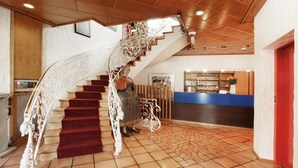 A grand staircase with ornate white railings and red carpeted steps leading to a reception area with a blue counter and a wooden bookshelf. - Hotel Schwarzwälder Hof (Freiburg im Breisgau)
