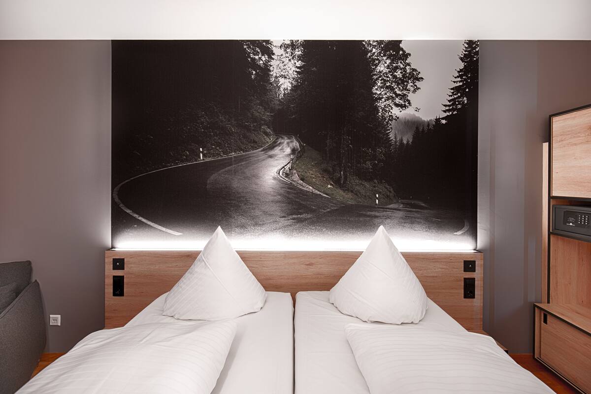 A hotel room with a large wall-mounted artwork of a winding road through a forest, a wooden headboard, two beds with white linens, and a small bedside table.