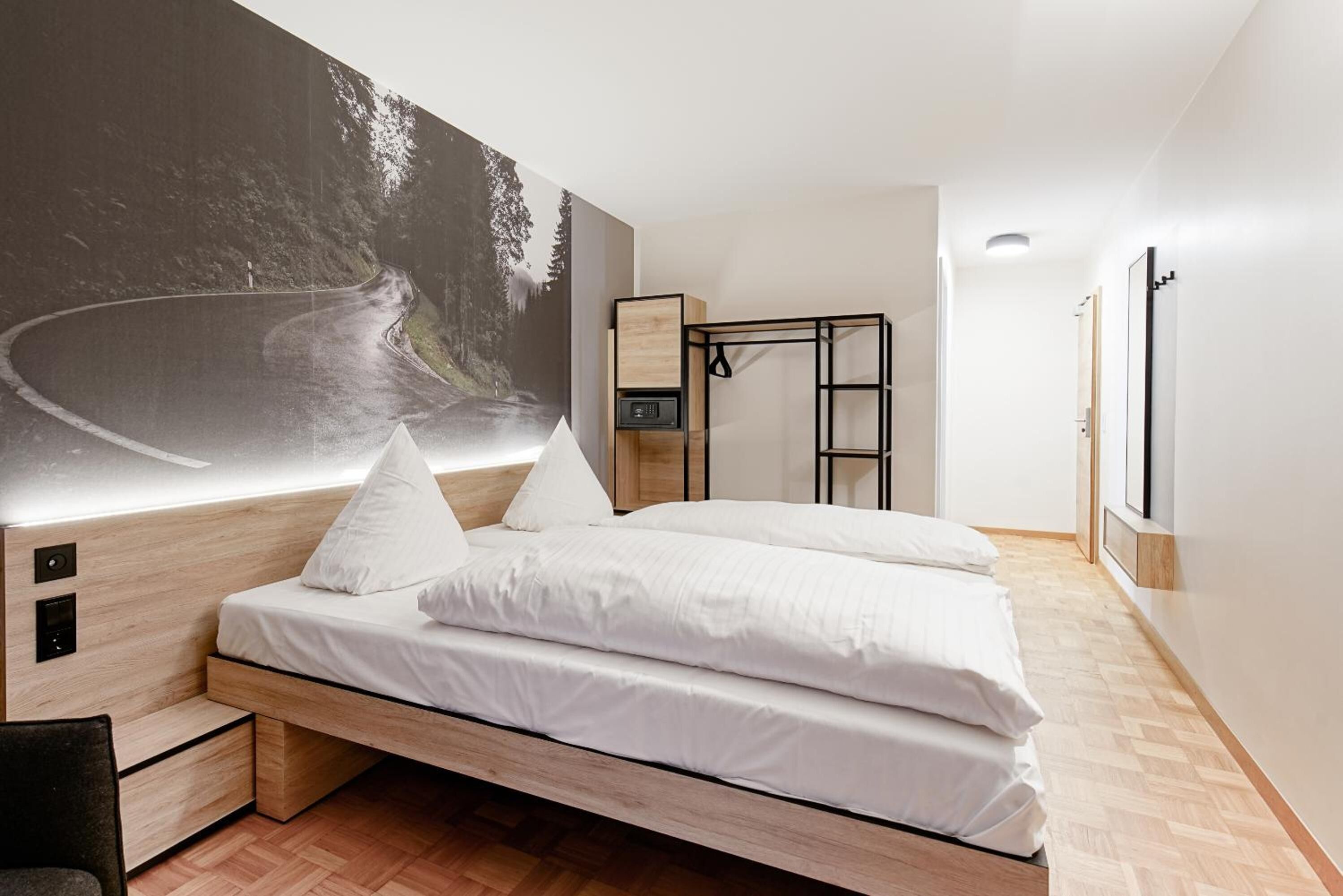 Ein modernes Schlafzimmer mit einem großen Bett, einem Kopfteil mit integrierten Bedienelementen, einem Nachttisch und einem großformatigen Wandbild, das eine kurvenreiche Straße zeigt.