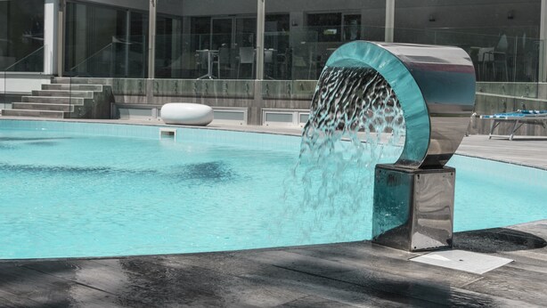 A modern pool with a water feature, surrounded by a glass railing and steps leading to a building with large windows. - Homeholiday Giulivo (Cellole)