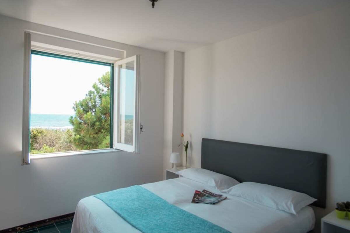 A bedroom with a large window, a bed with a grey headboard, a bedside table with a lamp, and a magazine on the bed.