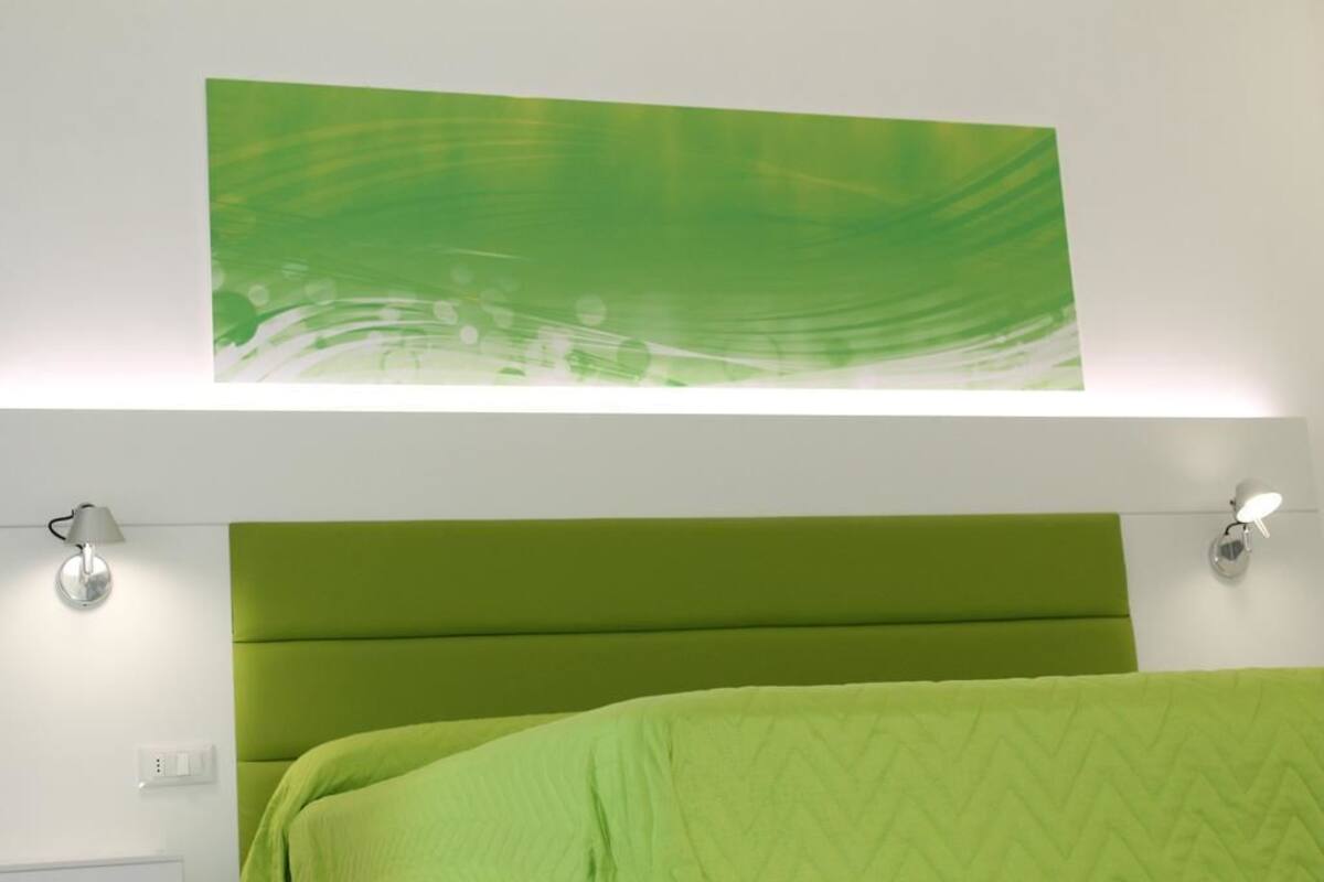 Ein Bett mit dunkelgrüner Kopfteil und Bettdecke, eine Wandleuchte und ein abstraktes Gemälde in Grün über dem Bett.