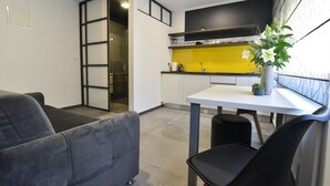 A modern kitchen with a yellow backsplash, a dining area with a white table and black chairs, and a grey sofa in a living area. - Mono Apartments & Rooms (Zadar)