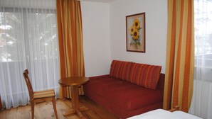 Une pièce avec un plancher en bois, un canapé rouge, une petite table ronde, une chaise en bois et un tableau encadré représentant des tournesols accroché au mur.