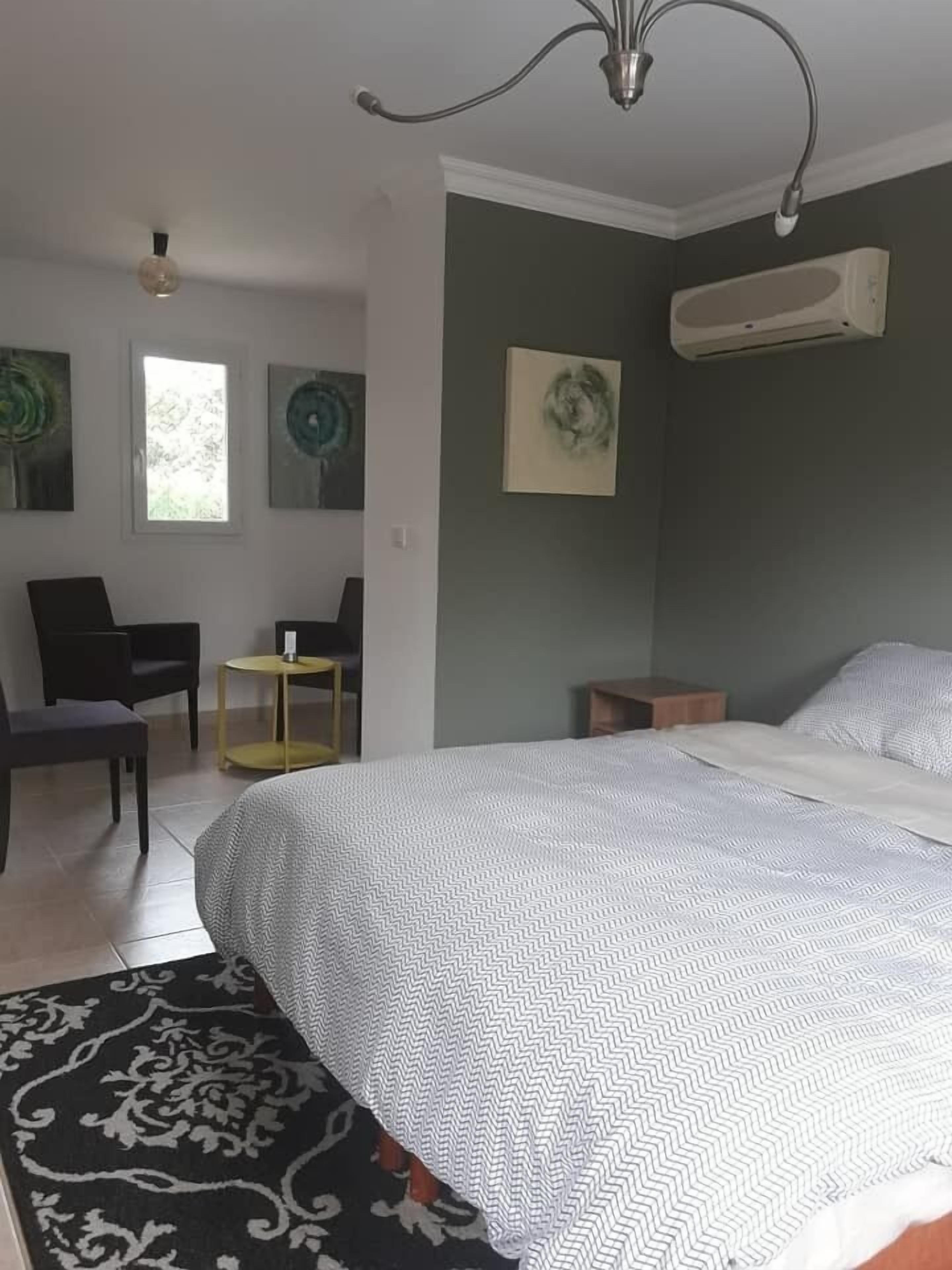 A bedroom with a bed, a desk, a chair, and a small table. There is a window, a wall-mounted air conditioner, and artwork on the walls.
