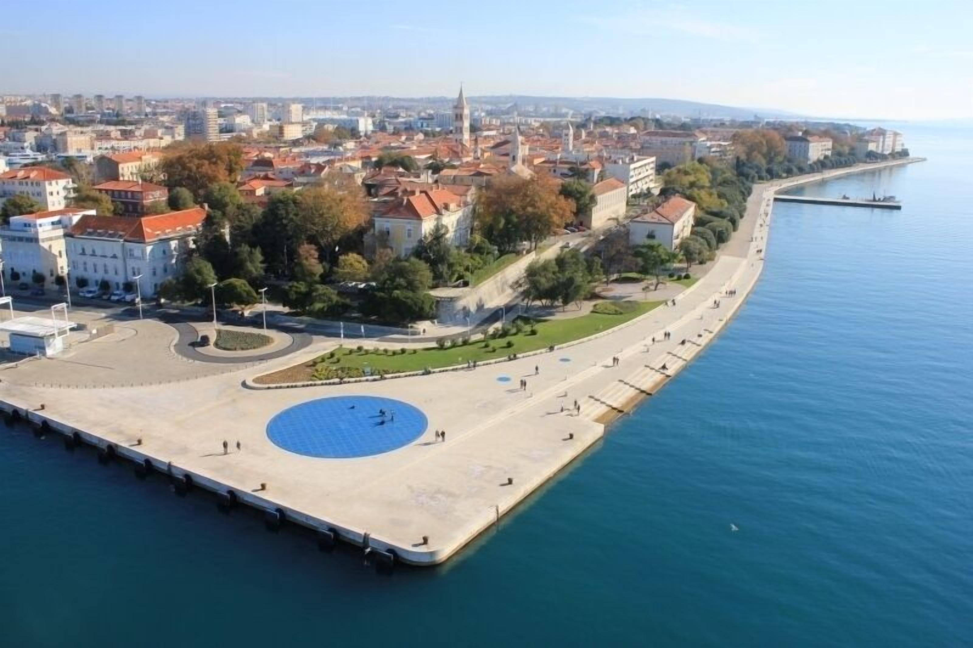Eine Luftaufnahme einer Küstenstadt mit einer markanten Uferpromenade, einem großen Platz und historischen Gebäuden mit roten Ziegeldächern.
