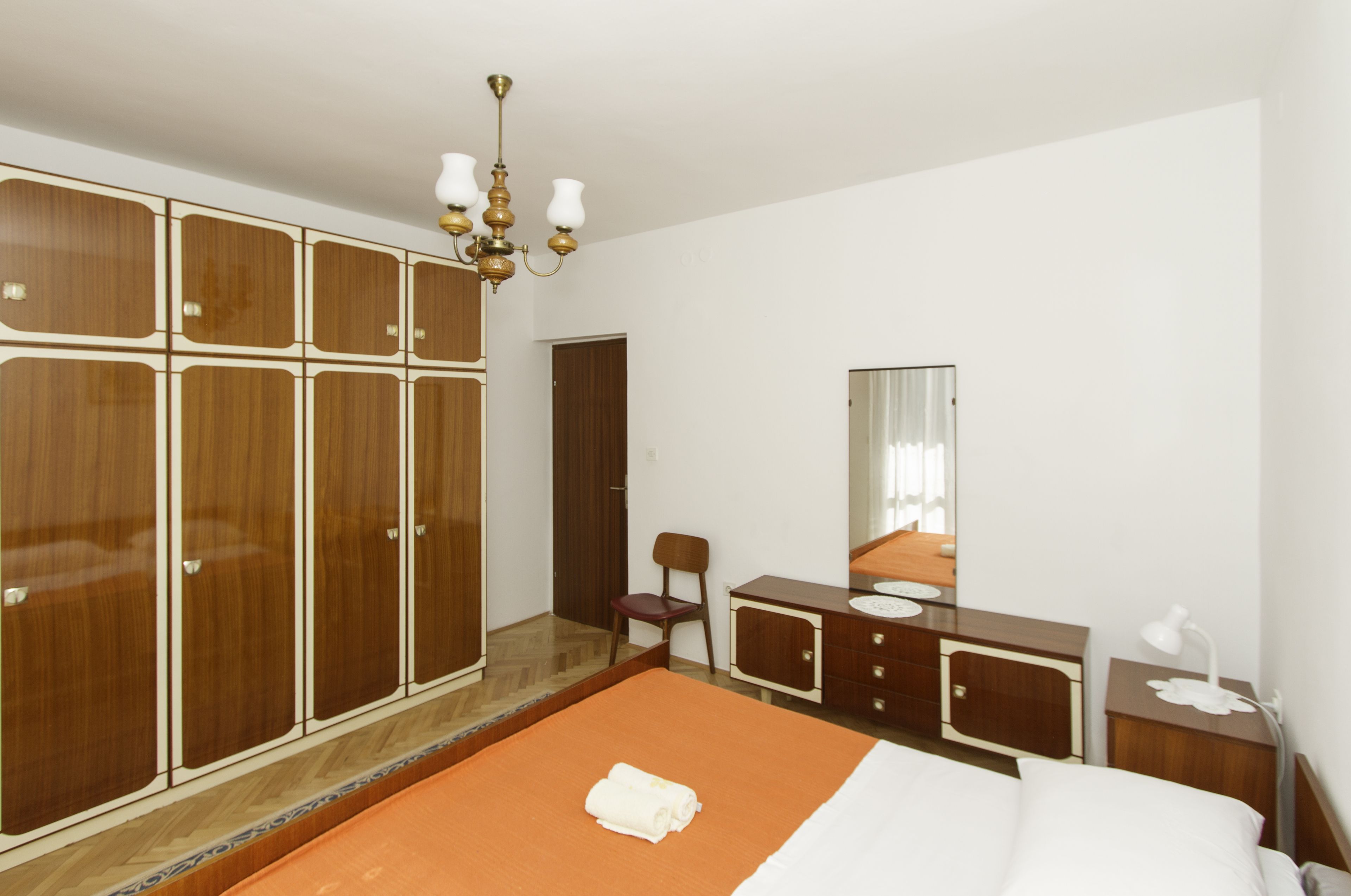 A bedroom with a large wardrobe, a bed with an orange bedspread, a bedside table with a lamp, and a mirror on the wall.
