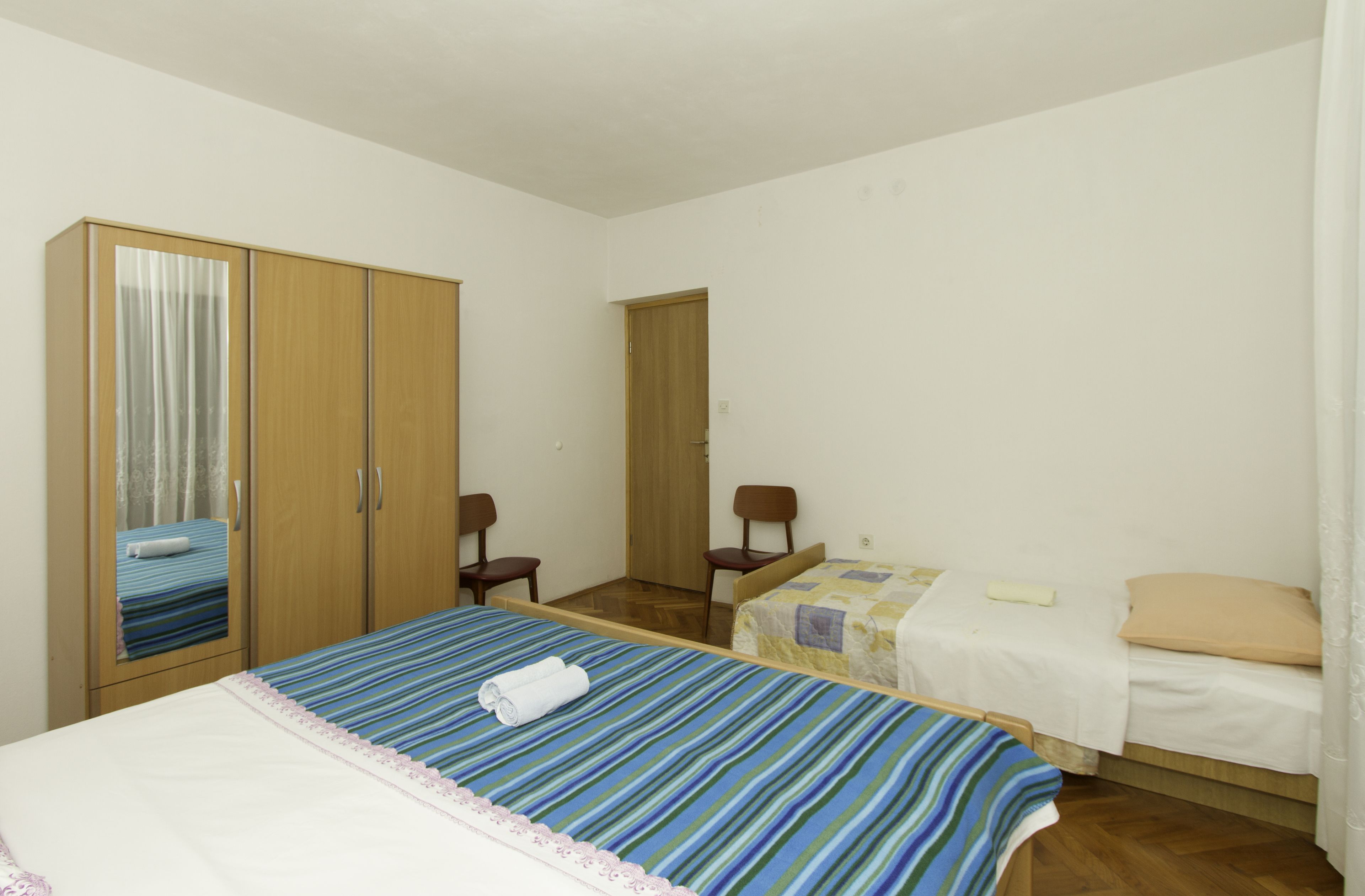 Een slaapkamer met een grote kast, een bed met een gestreept dekbed en een kleiner bed met een wit dekbed, plus een stoel.