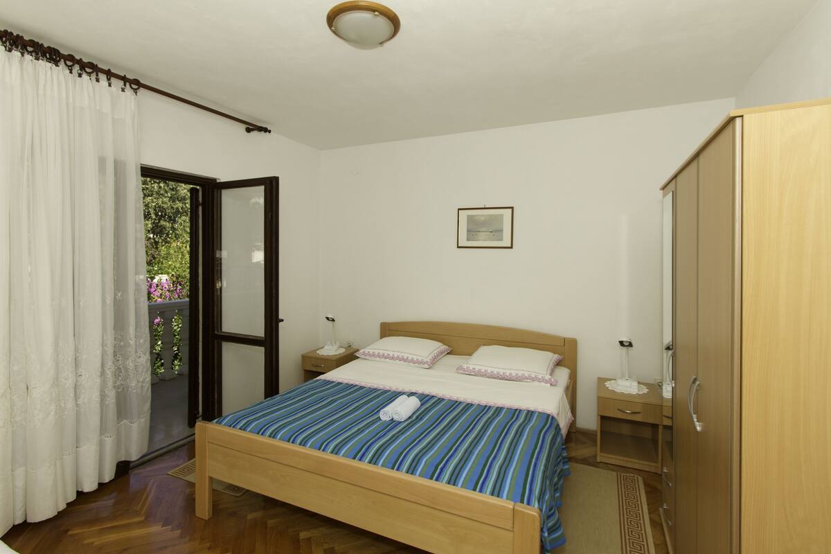 A bedroom with a wooden bed, white curtains, a small bedside table, a nightstand, a wardrobe, and a framed picture on the wall.