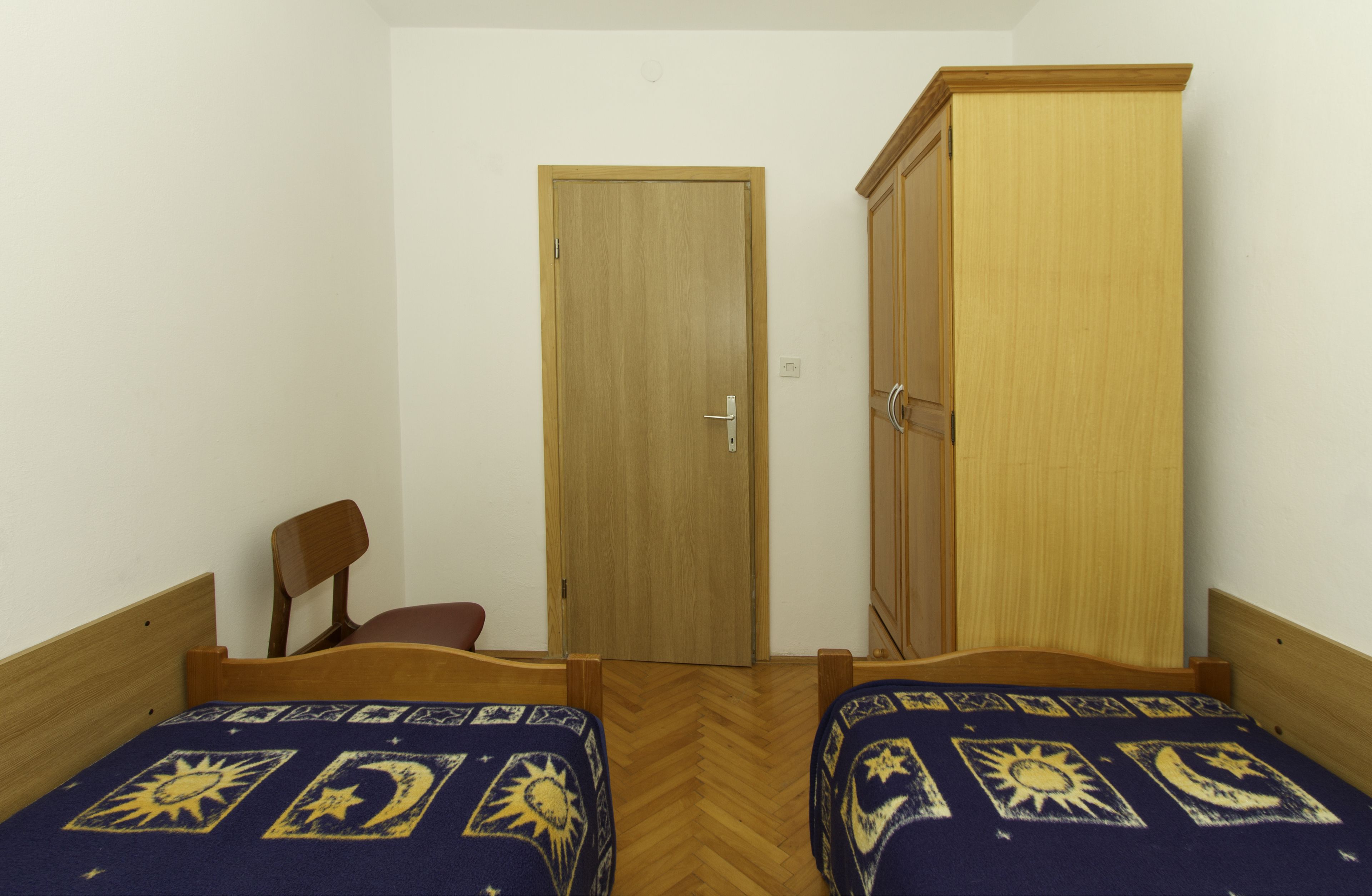 A room with two wooden beds with patterned bedding, a wooden wardrobe, a wooden chair, and a wooden door.