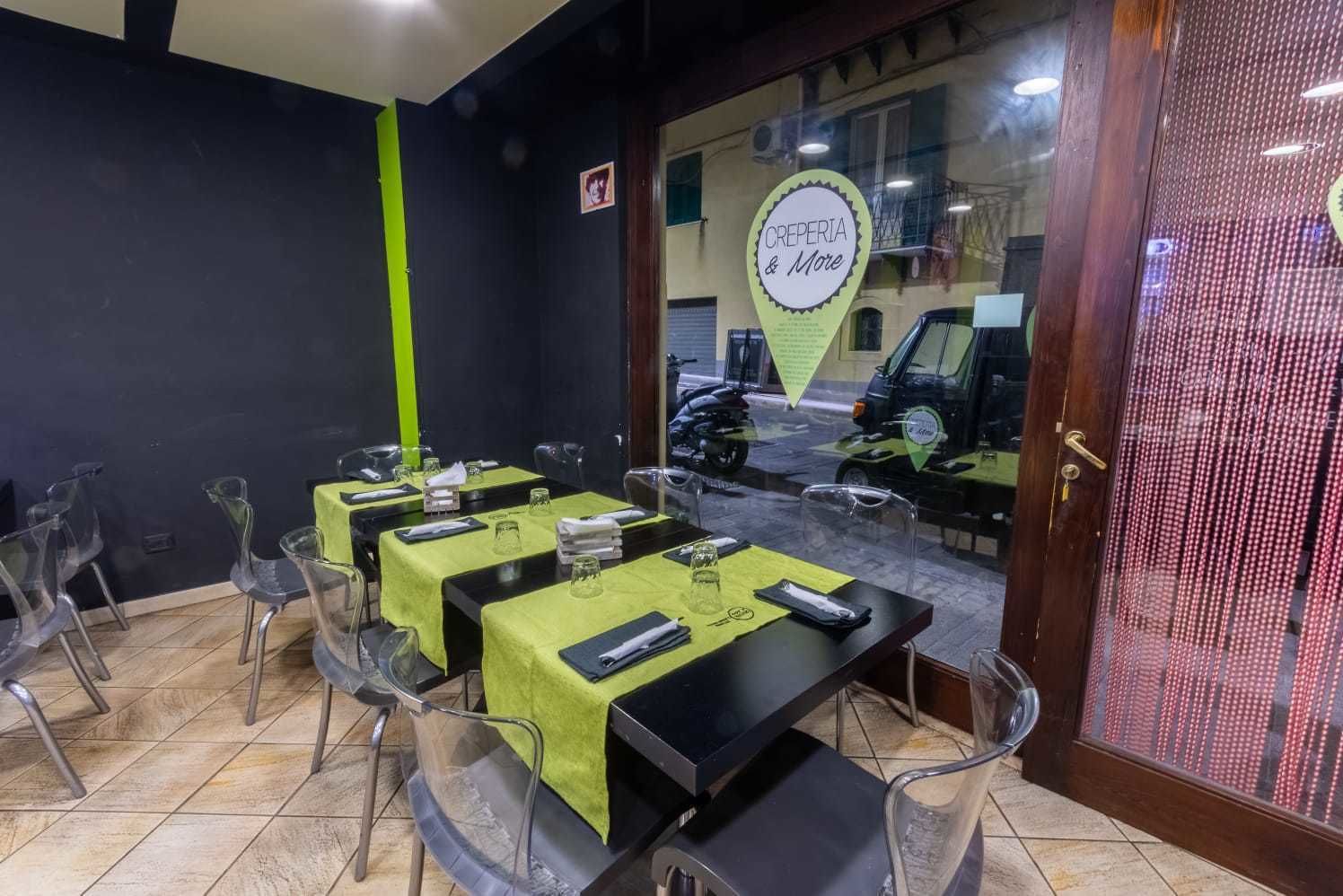 Una sala ristorante con tavoli apparecchiati, sedie trasparenti e un cartello che indica 'CREPERIA & More'.