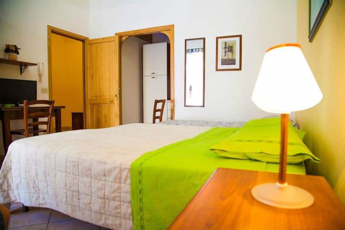A hotel room with a bed, a desk, a chair, a lamp, and a door leading to another room.