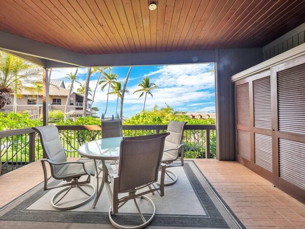 A covered outdoor area with a round table and chairs, a view of palm trees and a building, and a wooden ceiling.