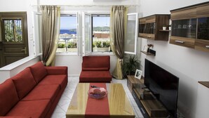 Sala de estar moderna com sofá vermelho, mesa de centro de madeira, televisão e vista para o mar através de grandes janelas.