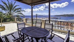 Uma área de estar coberta ao ar livre, com mesa e cadeiras, com vista para uma cidade costeira e o mar.