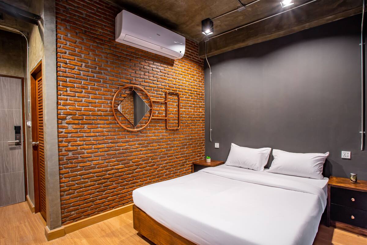 A modern bedroom with a brick wall, a circular mirror, a bed, and a nightstand with a lamp.